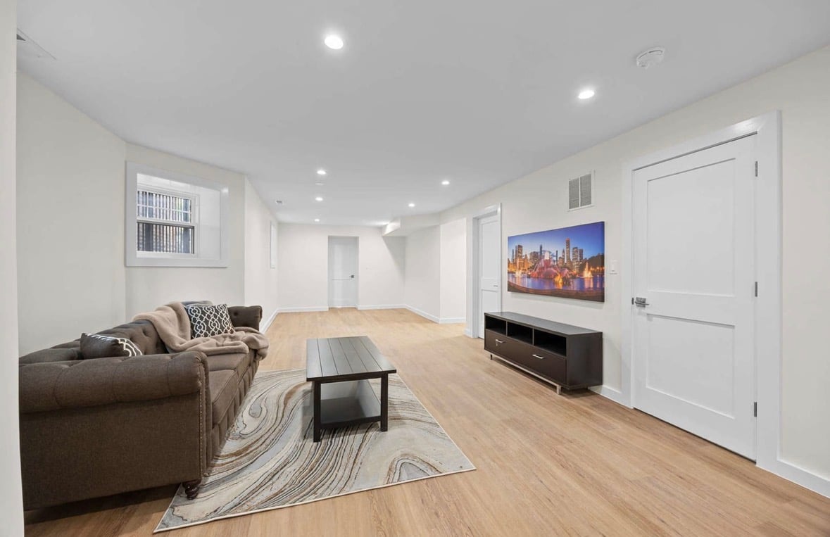 Bright basement living room with gray sofa, cityscape artwork, and wooden floor